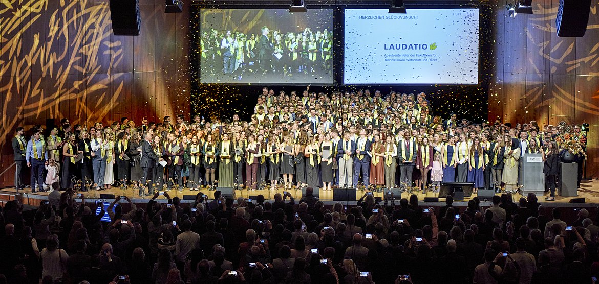 400 Absolventinnen und Absolventen jubeln bei ihrer Feier auf der Bühne, während Goldkonfetti auf sie regnet.