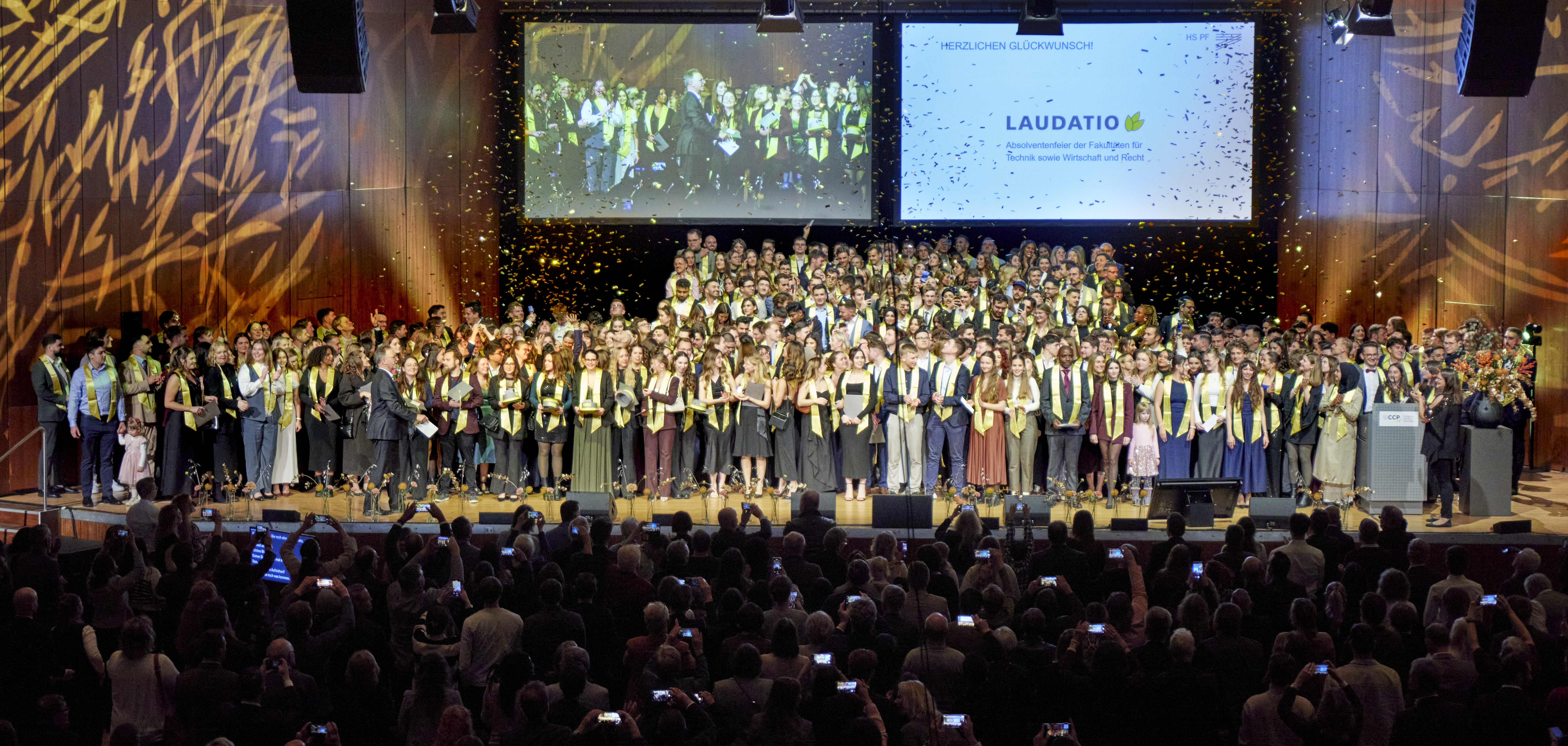 400 Absolventinnen und Absolventen jubeln bei ihrer Feier auf der Bühne, während Goldkonfetti auf sie regnet.